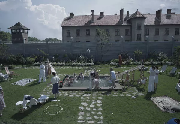 Szene aus dem Film The Zone of interest. Im Vordergrund ein großer Garten mit Steinwegen und einem kleinen Swimmingpool in der Mitte in dem Kinder spielen. Im Hintergrund eine Mauer und dahinter das Konzentrationslager Auschwitz.